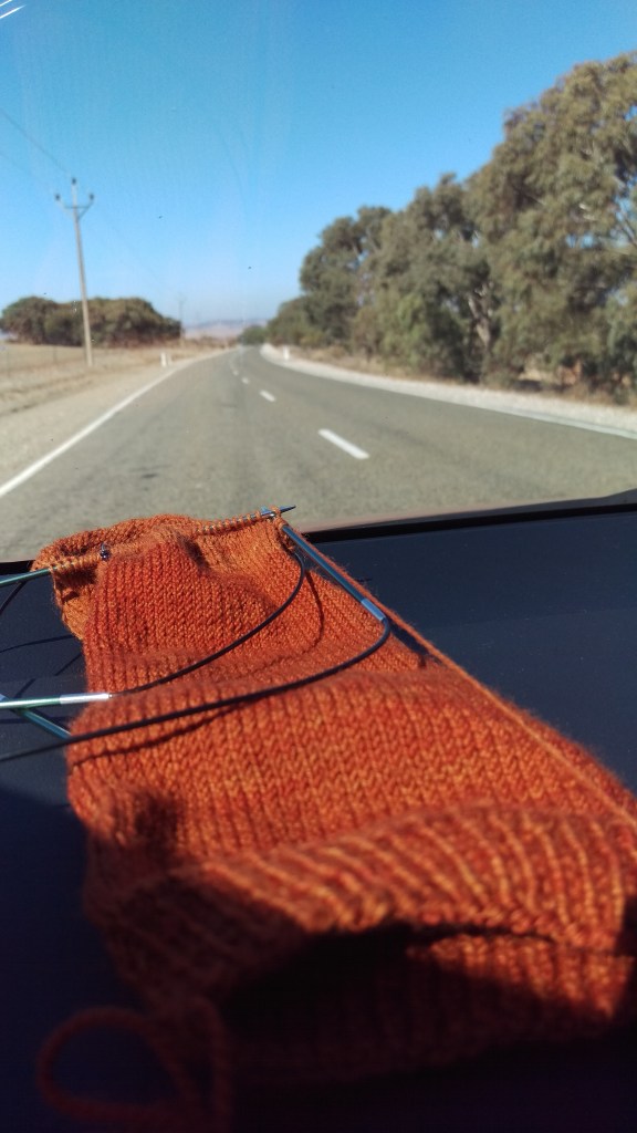 An orange sock in progress on the dashboard of a car, with a road and dry South Australian landscape and blue sky out of focus in the background.